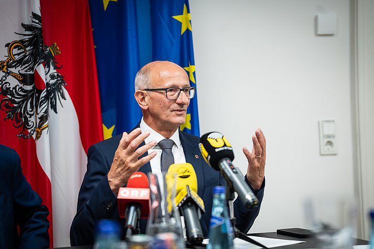 © Land tirol/Fitsch Mattle redend; im Hintergrund Fahnen, vor ihm auf dem Tisch Mikrofone