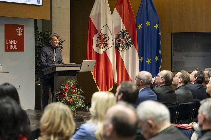 Ein Mann steht an einem Rednerpult, vor ihm sitzen mehrere Leute im Saal. 