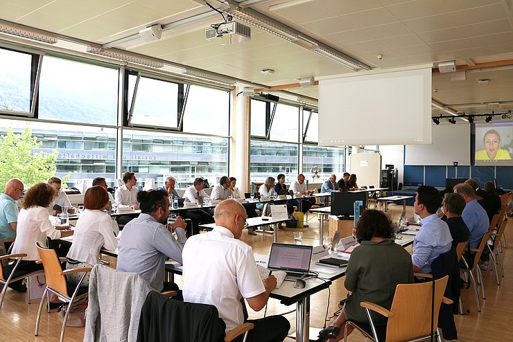 Konferenzsaal mit Personen an U-förmigem Tisch, auf dem Bildschirm die Ministerin