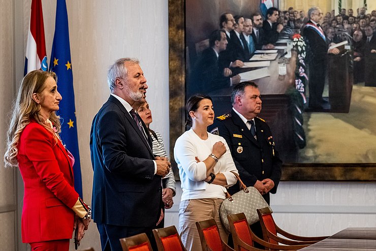 Mair, Sanader, eine Frau im roten Hosenanzug und ein kroatischer Feuerwehrmann im Parlament. Im Hintergrund ein riesen Gemälde mit einer Szene aus dem Parlament