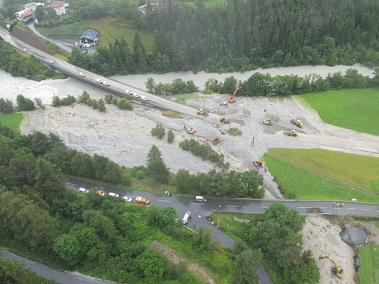 © Land Tirol/Drohnen-Einsatz-Gruppe Blick auf Fluss mit Brücke und einer weiteren Straße in einem Wald-Wiesen-Siedlungsgebiet. Bagger, Einsatzfahrzeuge sowie Autos sind zu sehen. Brücke ist teilweise überflutet. Unten und in der Mitte sind Ablagerungen eines Murgangs zu sehen.