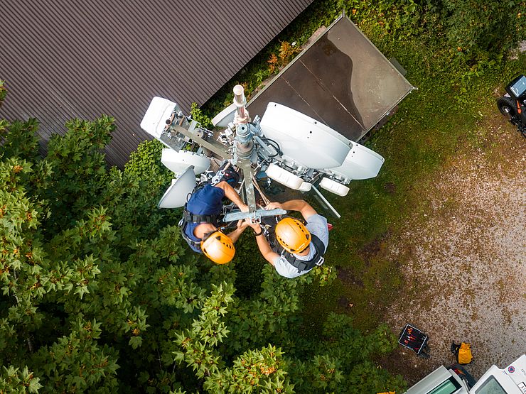 Aufnahme von der Spitze des Funkmasten (von oben mit Drohne aufgenommen); zwei Mitarbeiter an der Spitze und arbeiten
