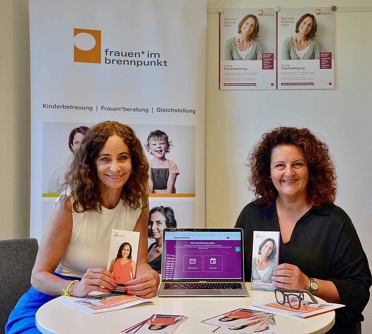 LRin und GFin halten Folder hoch, in der Mitte Laptop, auf dem die Seite der Online Frauenberatung offen ist