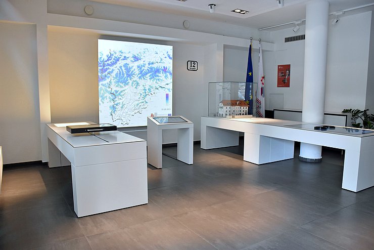 Auf dem Bild ist die Ausstellung zu sehen. Man kann drei weiße Tische mit Bildschirmen und im Hintergrund eine Karte der Europaregion Tirol-Südtirol-Trentino erkennen. 