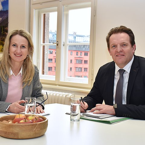 zwei Personen sitzen am Tisch, Blick in die Kamera