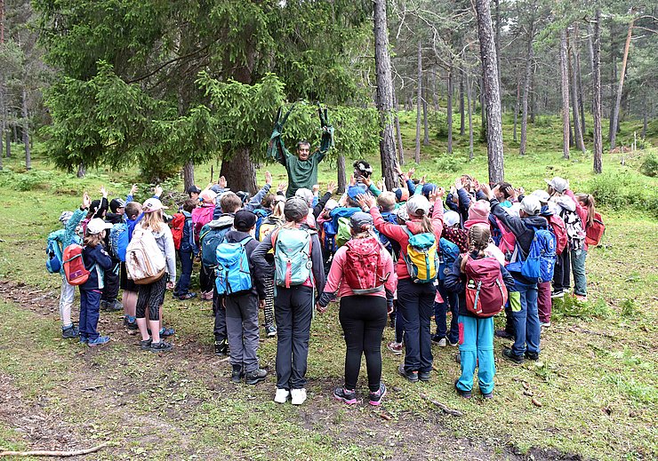 Eine große Gruppe an Kindern stehen im Wald und beobachten ein Theaterspiel.