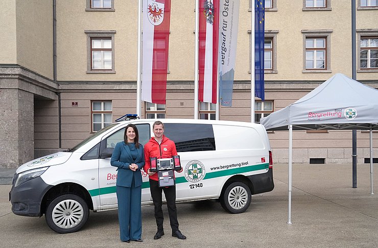 Mair und Wimmer vor Auto und neben Zelt stehend mit Defi in der Hand