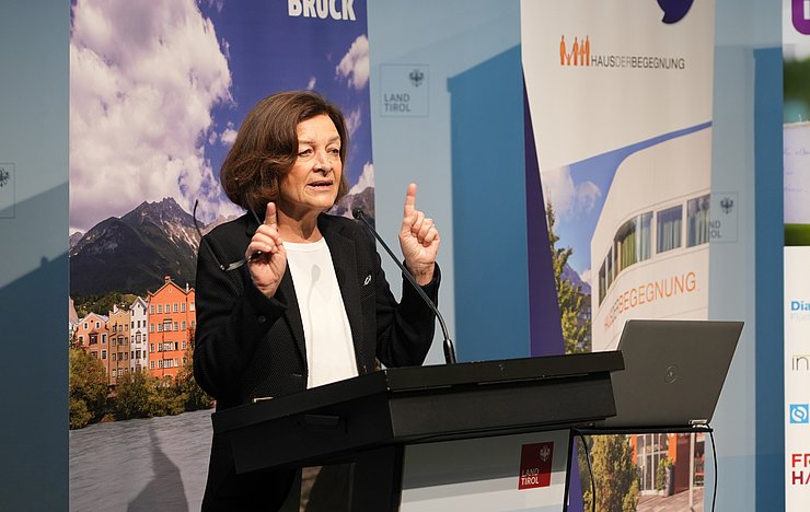 © Land Tirol/Saxl Karin Gutierrez-Lobos mit kurzem braunem Haar und schwarzem Blazer spricht am Rednerpult und hebt dabei beide Hände. Im Hintergrund sind Banner des Landes Tirol, der Stadt Innsbruck und des Hauses der Begegnung zu sehen.