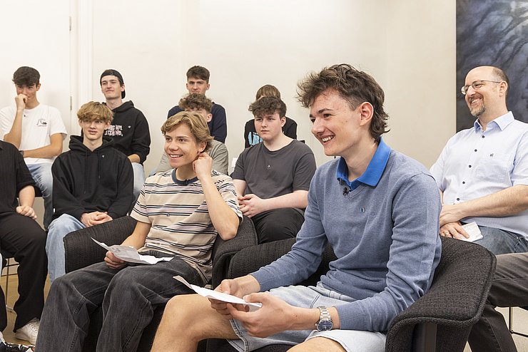 Zwei Schüler sitzen vor der restlichen Klasse, halten Karten und lachen.