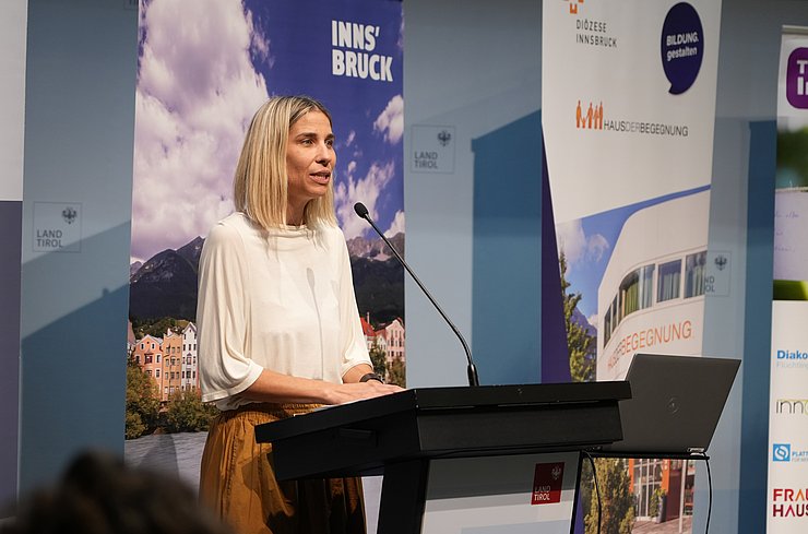 © Land Tirol/Saxl Janine Bex mit schulterlangen blonden Haaren und heller Bluse spricht am Rednerpult. Hinter ihr stehen Banner der Stadt Innsbruck, des Landes Tirol und des Hauses der Begegnung.