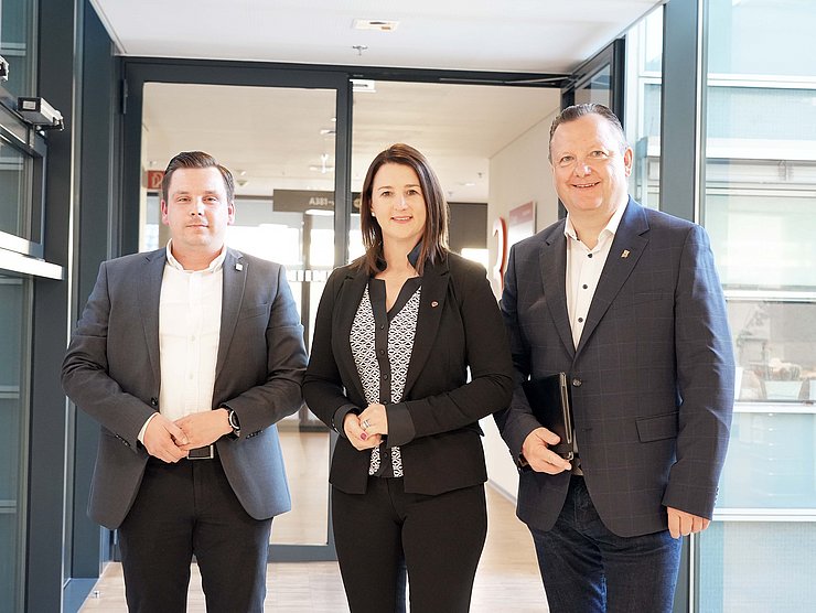 Gruppenfoto mit einer Frau in der Mitte, die von zwei Herren umringt wird.