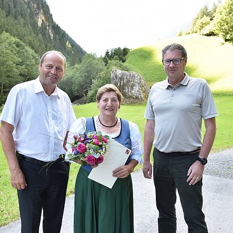 Gruppenfoto von Landeshauptmannstellvertreter Josef Geisler, Martha Auer und Klaus Niedertscheider. Im Hintergrund ist eine Wiese, Bäume und Berghang zu sehen