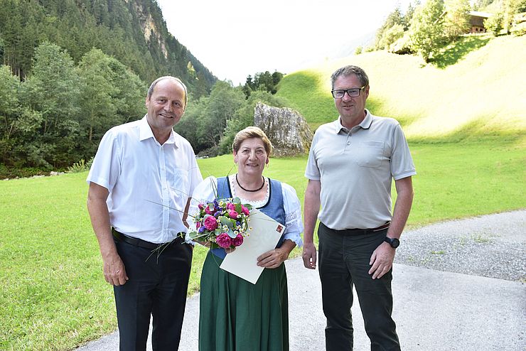 Gruppenfoto von Landeshauptmannstellvertreter Josef Geisler, Martha Auer und Klaus Niedertscheider. Im Hintergrund ist eine Wiese, Bäume und Berghang zu sehen