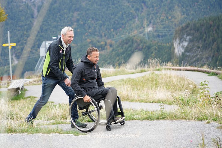 © Land Tirol/Brandhuber Mann schiebt anderen Mann im Rollstuhl auf Wanderweg