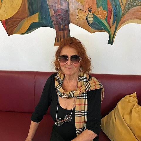 Marion Fischer trägt eine Sonnenbrille und sitz auf einem roten Ledersofa mit einem gelben Kissen..