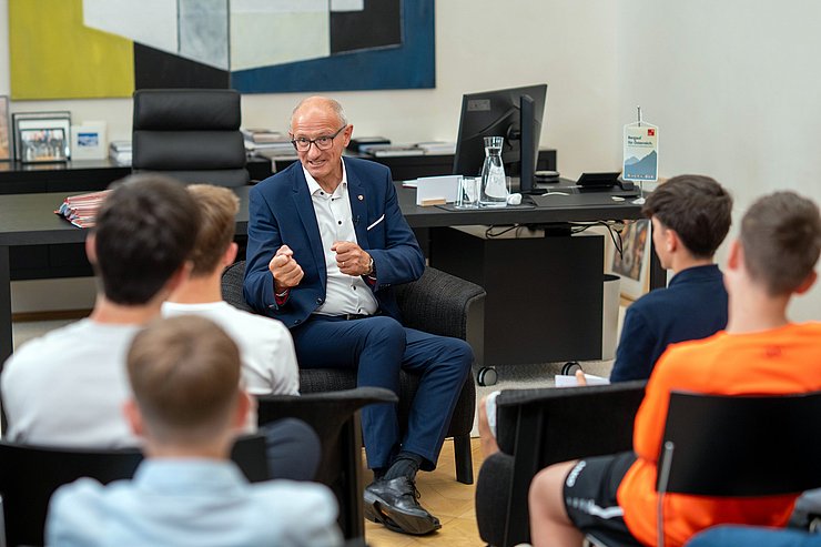 © Land Tirol/Jovanovic Mann im Anzug sitzt SchülerInnen gegenüber und spricht.