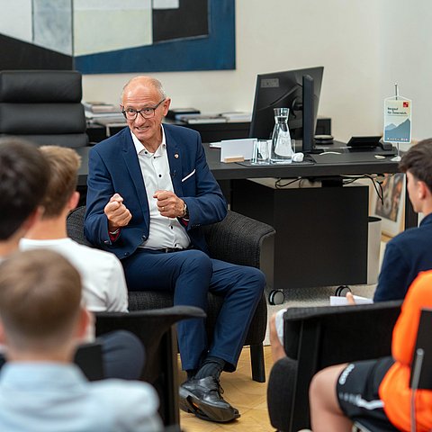 Mann im Anzug sitzt SchülerInnen gegenüber und spricht.