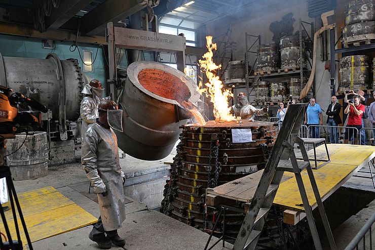 Erhitztes Metall wird in Form gegossen, drumherum Menschen in Schutzausrüstung und Schaulustige