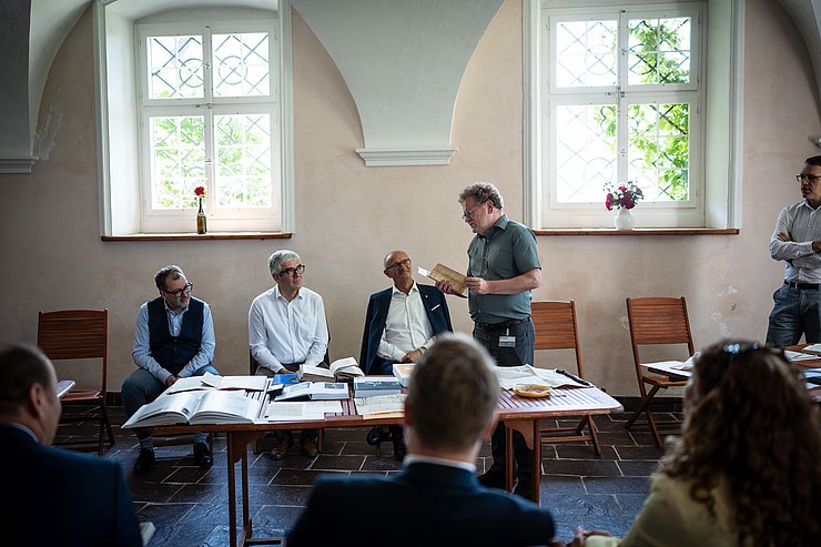 Person liest aus Buch vor, steht vor Tisch mit Dokumenten