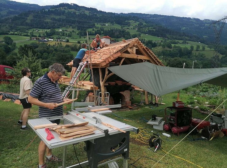 Mehrere Personen arbeiten an dem Schindeldach. Zwei Personen befinden sich auf dem Dach und befestigen Holzschindeln, eine Leiter lehnt am Dachrand. Im Vordergrund steht eine Person an einer Tischkreissäge mit Holzleisten. Um den Bau herum liegen Werkzeuge, Maschinen, Kabel und Holzmaterial. 