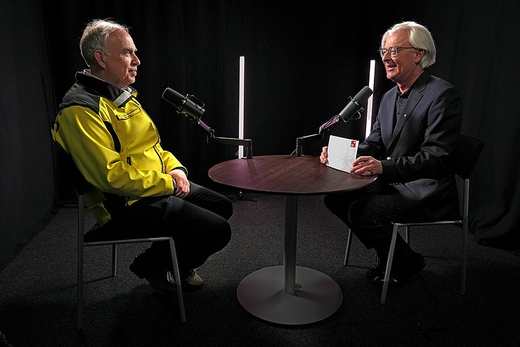 Zwei Männer sitzen an einem Tisch mit Mikrofonen und dunklem Hintergrund