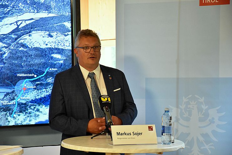 Bürgermeister Markus Sojer steht vor einem weißen Tisch mit Mikrofon. Im Hintergrund ist eine Landkarte der Region zu sehen.