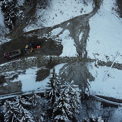 Drohnenaufnahme der B 171: Eine Mure geht über die gesamte Straße und verdeckt diese; ein Bagger steht auf der Mure und schaufelt Material in einen LKW