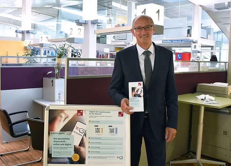Landesrat Anton Mattle hält Informationsbroschüre zur Handy-Signatur bzw. ID Austria in der Hand, daneben ein A-Steher mit Informationsblättern