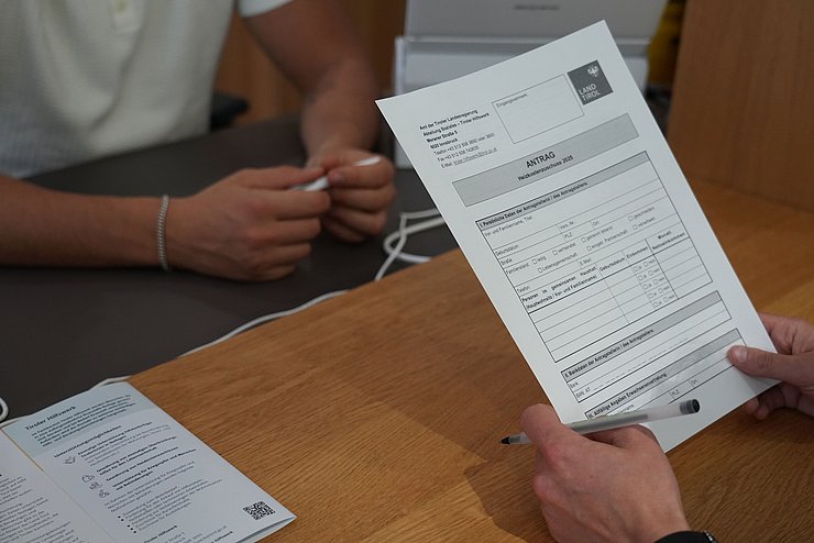 zwei Personen sitzen am Tisch, eine hält ein Antragsformular 