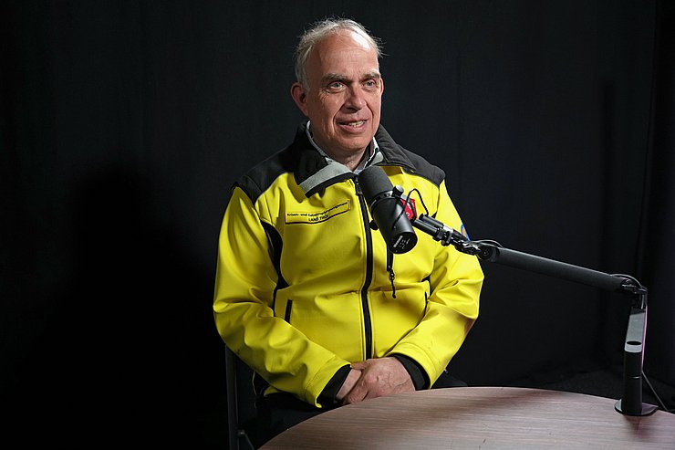 Mann mit gelber Jacke vor dunklem Hintergrund mit Podcastmikrofon