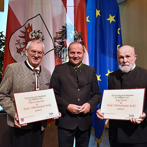 Landeshauptmann-Stellvertreter Josef Geisler steht zwischen den Preisträger Helmut Jenewein und Josef Zeisler vor der europäischen, österreichischen und Tiroler Flagge