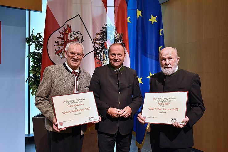 Landeshauptmann-Stellvertreter Josef Geisler steht zwischen den Preisträger Helmut Jenewein und Josef Zeisler vor der europäischen, österreichischen und Tiroler Flagge