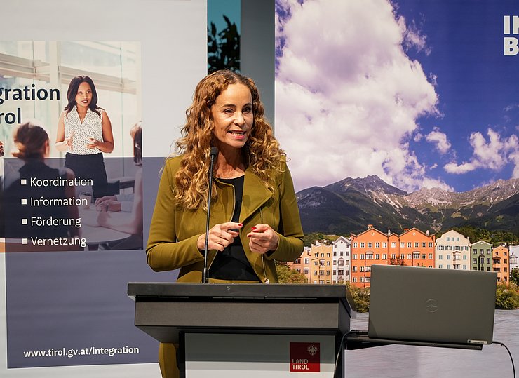 © Land Tirol/Saxl Eva Pawlata in olivgrünem Anzug spricht am Rednerpult mit Land-Tirol-Logo. Im Hintergrund sind ein Informationsbanner zur Integration in Tirol und ein Banner mit Innsbrucker Altstadt-Panorama zu sehen.