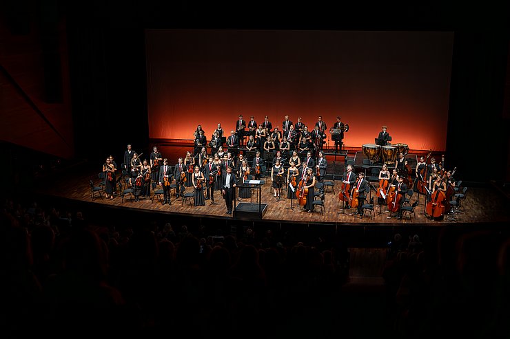 Die Mitglieder eines Orchesters stehen und blicken Richtung Publikum.