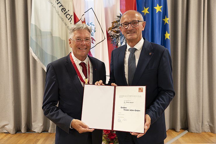 © Land Tirol/Sedlak Zwei Männer stehen vor Fahnen und blicken in die Kamera.