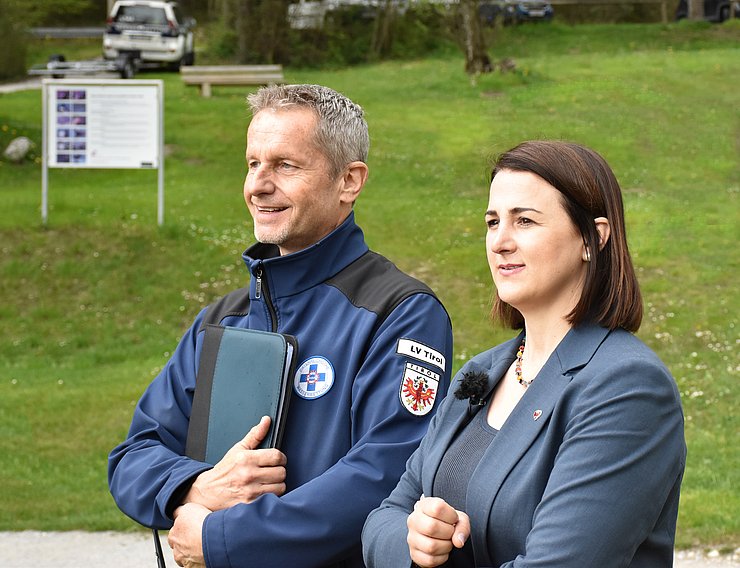 Stock (links) und Mair stehen nebeneeinander am Ufer. Beide lächeln. Stock hat eine balue Jacke der Wasserrettung an und hält eine Mappe fest. Mair hat einen blauen Blazer. Im Hintergrund eine grüne Wiese und ein Auto der Wasserrettung mit einem Bootsanhänger