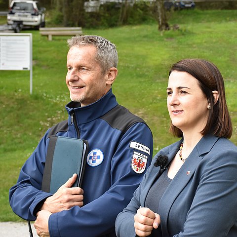 Stock (links) und Mair stehen nebeneeinander am Ufer. Beide lächeln. Stock hat eine balue Jacke der Wasserrettung an und hält eine Mappe fest. Mair hat einen blauen Blazer. Im Hintergrund eine grüne Wiese und ein Auto der Wasserrettung mit einem Bootsanhänger