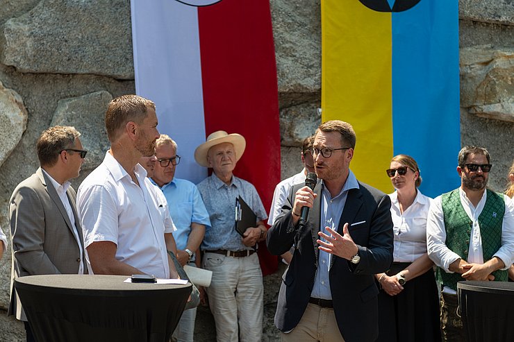 zwei Männer stehen sich gegenüber, einer mit Mikrofon, im Hintergrund sind mehrere Personen zu sehen