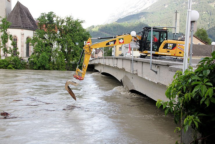 Fluss mit erhöhtem Wasserspiegel an dem ein Bagger von der Brücke aus arbeitet.