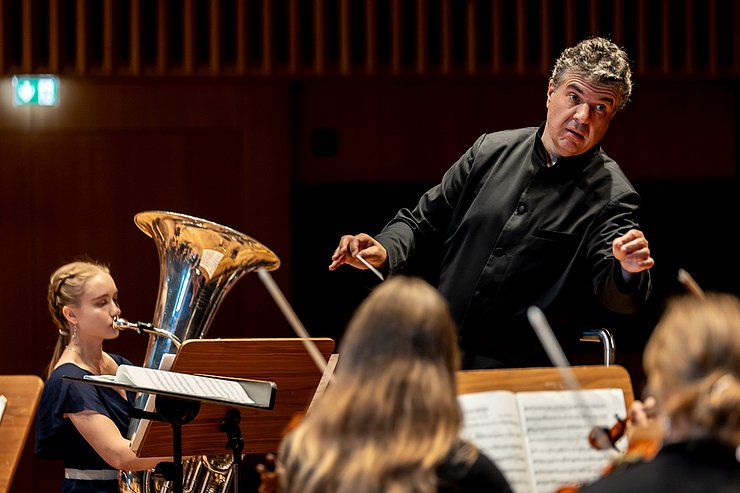 © Land Tirol/Sophie Kary Ein Mann dirigiert ein Orchester, Solistin ist eine Tubistin.
