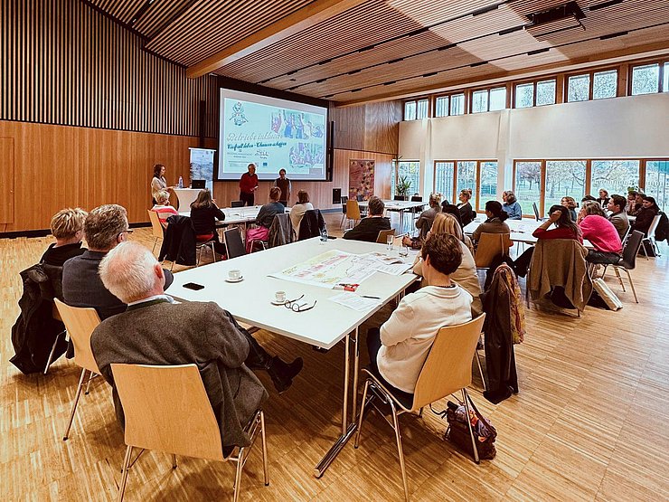 Mehrere Menschen sitzen an Tische und hören einer Präsentation zu.