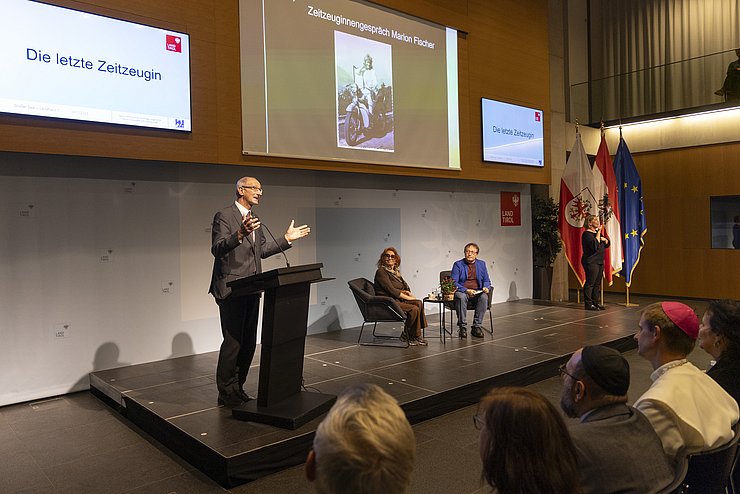 Am 9. November 2023 begrüßte LH Anton Mattle (stehend links) die Zeitzeugin Marion Fischer (sitzend Mitte) im Großen Saal im Landhaus, die vor Publikum ein Gespräch mit dem Zeithistoriker Horst Schreiber (sitzend rechts) von ERINNERN:AT führte. Ganz rechts steht auf dem Podium eine Gebärdendolmetsch.