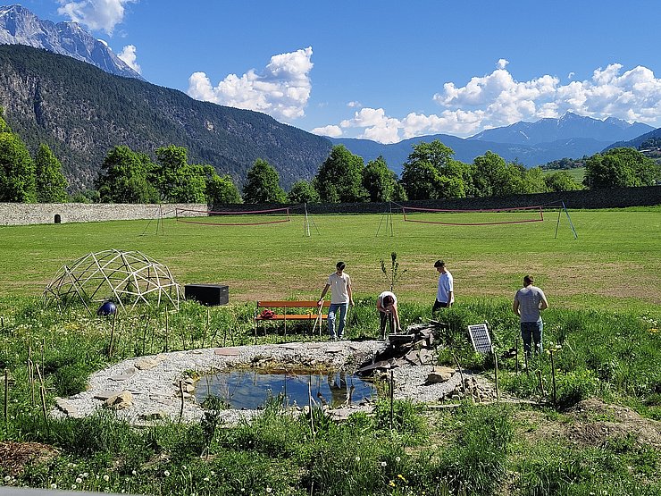 Ein naturnaher Schulgarten mit einem kleinen, runden Teich aus Steinen in der Mitte: Mehrere Jugendliche stehen am Rand des Teichs und arbeiten mit Werkzeugen. Umgeben ist der Teich von Wiesen, jungen Pflanzen und Sitzbänken. Im Hintergrund liegen Sportwiesen mit Netzen, Bäume, eine Steinmauer und eine weite alpine Berglandschaft unter blauem Himmel mit Wolken.