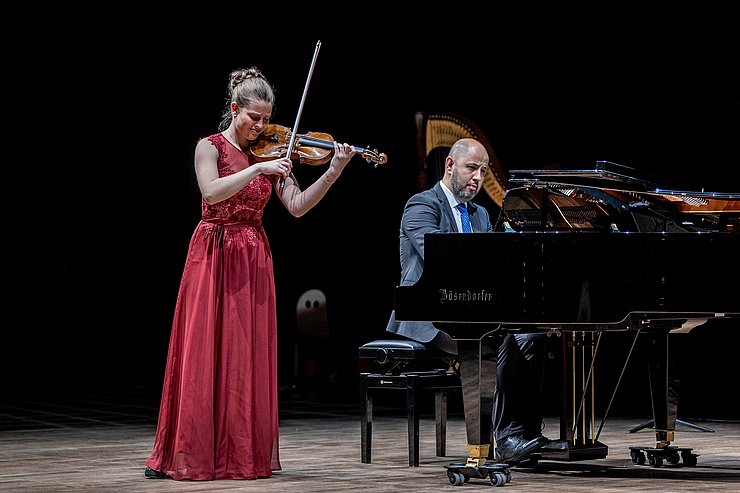 Ein Mädchen in rotem Kleid spielt Geige und wird von einem Mann am Flügel begleitet. 