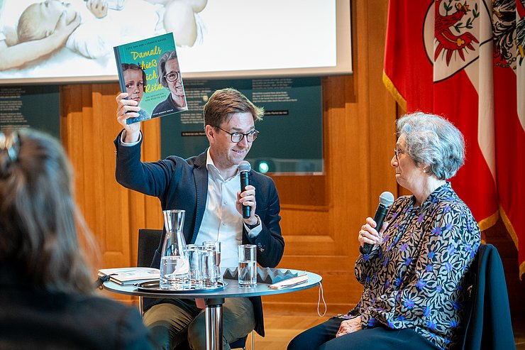 © Land Tirol/Neuner Der Moderator Siegele hält ein Buch in die Luft.