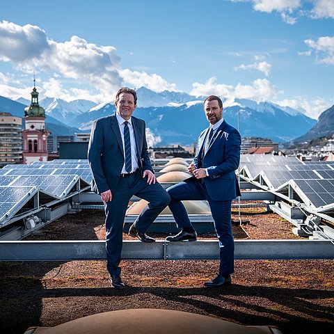 Zumtobel und Dornauer auf Dach Landhaus; im Hintergrund Innsbruck; am Dach PV-Anlagen; beide stehen mit jeweils einem Fuß auf Stahlträger der Anlage