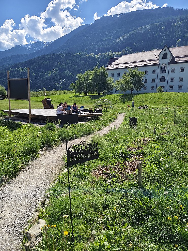 Ein sonniger Schulgarten in alpiner Umgebung: Ein schmaler Kiesweg führt durch eine blühende Wiese zu einer niedrigen Holzplattform, auf der mehrere Personen sitzen. Daneben wachsen Beete mit Wildblumen und Kräutern; ein kleines Metallschild mit der Aufschrift „Willkommen“ steht am Weg. Im Hintergrund sind grüne Berghänge, ein weißes Gebäude und ein blauer Himmel mit Wolken zu sehen.