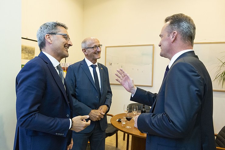 Mattle und Kompatscher im Gespärch mit Paasch; Paasch spricht gerade und hebt die Hand; im Hintergrund eine Wand mit Bildern darauf und ein Besprechungstisch 