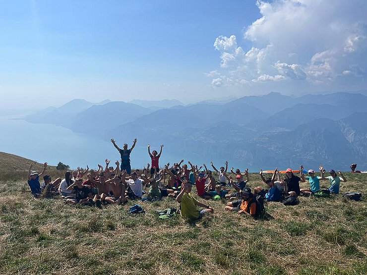 Gruppe Jugendlicher streckt Hände nach oben, sitzt auf Wiese - dahinter Bergkulisse und See