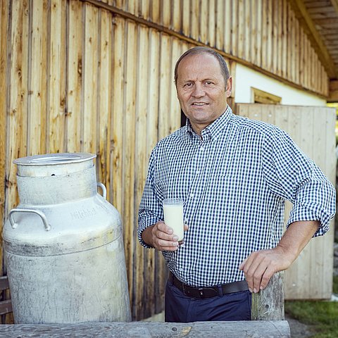 Josef Geisler steht vor einer Hütte und neben einer Milchkanne und hält ein Glas mit Milch in der Hand